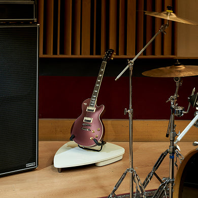 Burgundy electric guitar on white Aeropick stand in professional music studio setting with drum kit and amplifier